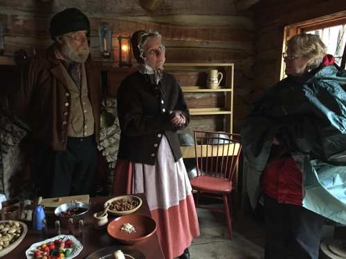 Three people in historical attire engage in conversation inside a rustic cabin, with a table of food items nearby.