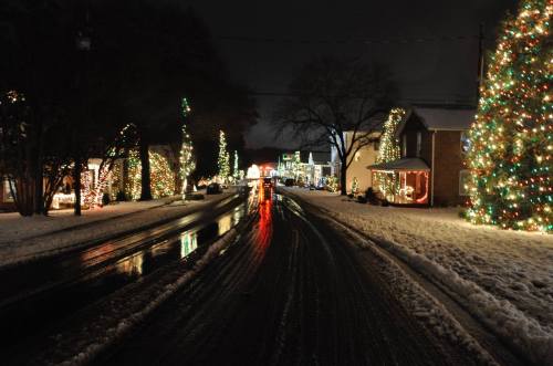 A snowy street at night, lined with houses decorated in colorful holiday lights reflecting on wet pavement.