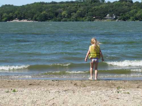 A child in a life jacket stands at the water's edge, looking out at the waves on a sunny day.