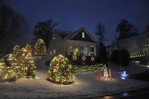 A cozy house decorated with colorful Christmas lights, surrounded by snow-covered trees and festive displays at dusk.