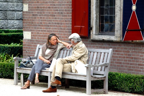 A woman and an older man sit on a bench, engaged in a conversation in a garden setting.