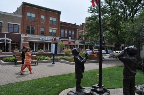 Two children statues salute a flagpole in a park, with shops and people walking in the background.
