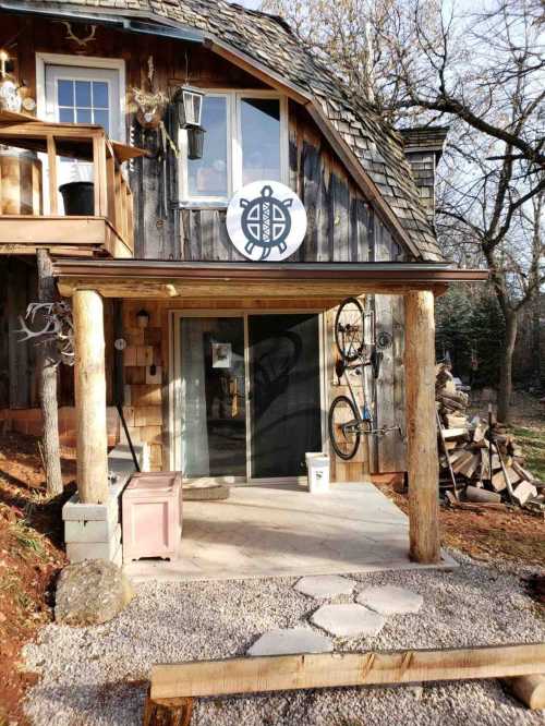 A rustic wooden cabin with a turtle logo, porch, and bicycle, surrounded by trees and gravel pathway.