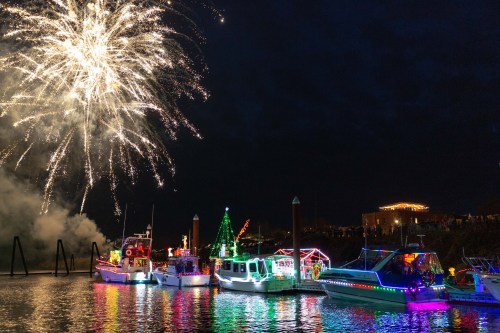 Colorful boats decorated with lights on a calm waterway, with fireworks bursting in the night sky above.