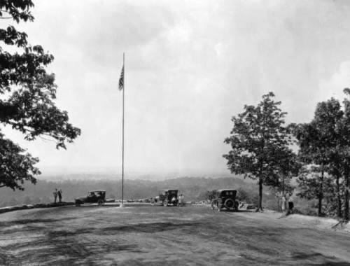 Historic black-and-white photo of cars parked near a flagpole, surrounded by trees and a scenic view.
