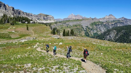A group of hikers walking along a trail in a mountainous landscape with green meadows and rocky peaks in the background.