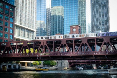 A train crosses a bridge over a river, surrounded by tall buildings and a clock tower in an urban setting.