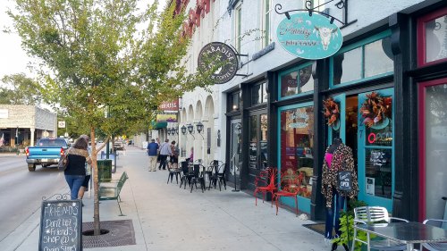 A charming street scene with shops, outdoor seating, and people walking along a tree-lined sidewalk.