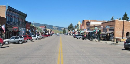 A sunny street view of a small town with shops, parked cars, and mountains in the background.