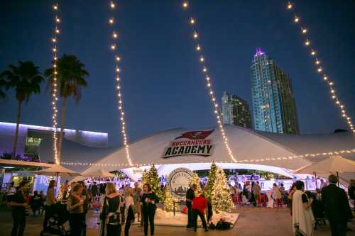 A festive outdoor event at the Buccaneers Academy, adorned with lights and holiday decorations, with city buildings in the background.