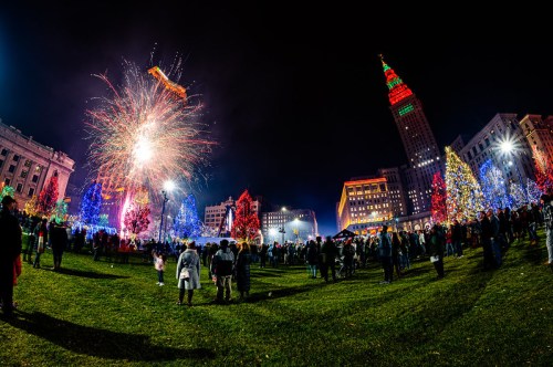 A festive scene with colorful lights, fireworks, and a crowd enjoying a holiday celebration in a city park at night.