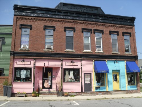 A charming brick building with a pink storefront and colorful blue and yellow shops, featuring flower planters outside.