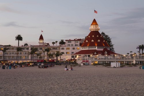 Historic hotel with a red-topped tower, palm trees, and beach at dusk, with people gathered on the sand.