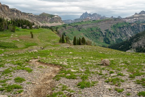 A winding dirt path leads through a lush green valley, surrounded by rocky mountains under a cloudy sky.