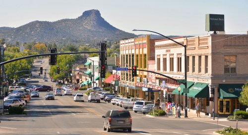 A bustling street scene with shops and cars, set against a backdrop of a mountain under a clear blue sky.