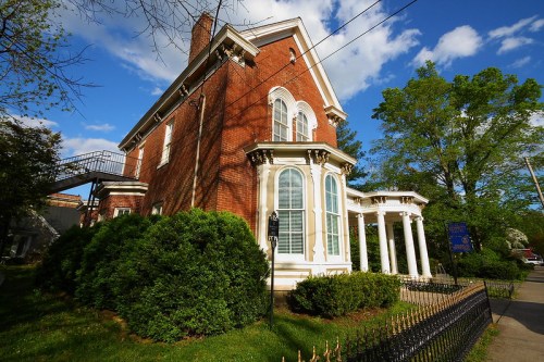 A historic brick house with large windows, white columns, and lush greenery, set against a blue sky with clouds.