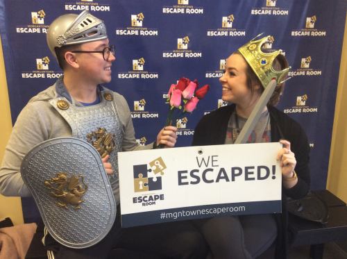 A man in armor and a woman in a crown hold roses and a sign that says "We Escaped!" in an escape room setting.