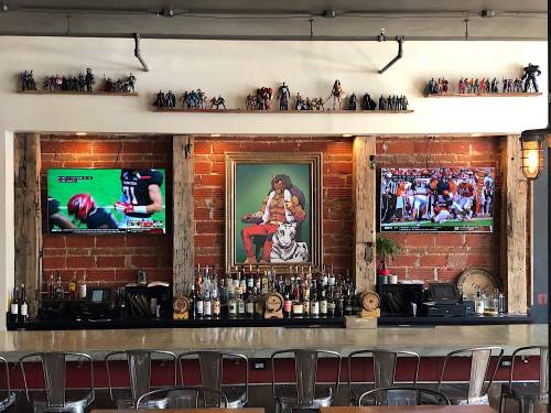 A bar with two TVs showing sports, a framed painting, and shelves displaying figurines against a brick wall.