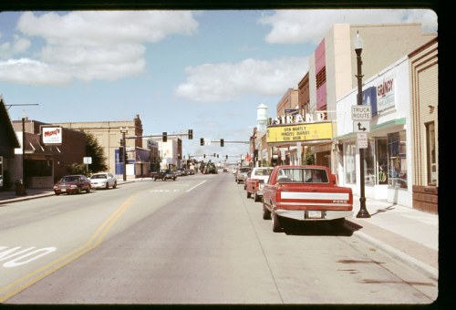 A street view featuring shops, parked red trucks, and traffic lights under a blue sky with clouds.