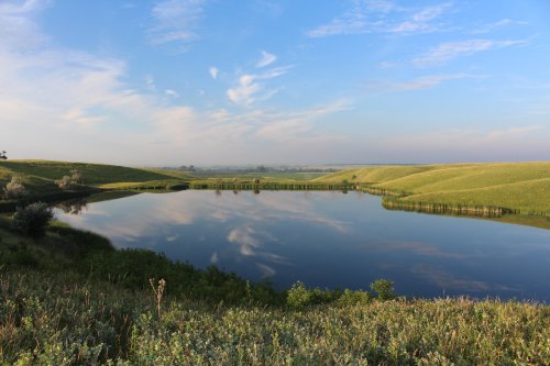 A serene landscape featuring a calm lake surrounded by green hills and a clear blue sky with soft clouds.