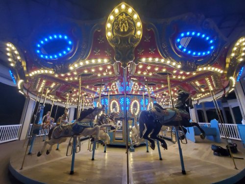 A brightly lit carousel with ornate horses, illuminated by blue and white lights, set in a nighttime scene.