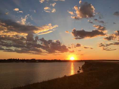 A serene sunset over a calm lake, with colorful clouds reflecting in the water and a grassy shoreline.