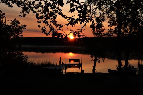 A serene sunset over a calm lake, framed by trees and a silhouette of a dock.