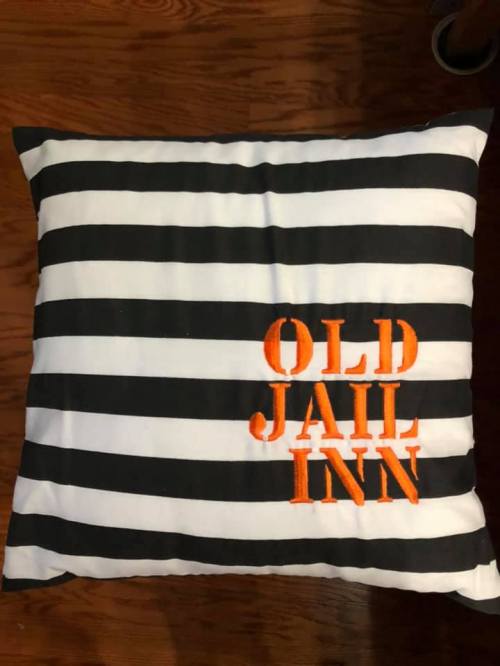 A square pillow with black and white stripes featuring the text "OLD JAIL INN" in orange embroidery.