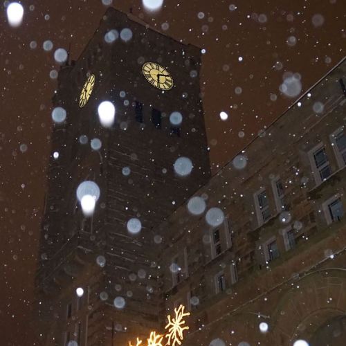 A clock tower illuminated at night, surrounded by falling snowflakes and a warm glow from nearby lights.