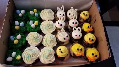 A box of colorful cupcakes decorated as bunnies, chicks, and Easter eggs on green grass.