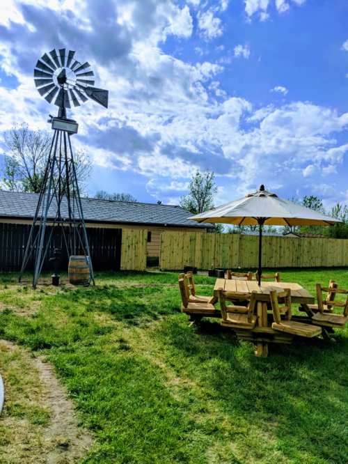 A grassy outdoor area featuring a windmill, picnic table, and umbrella under a partly cloudy sky.