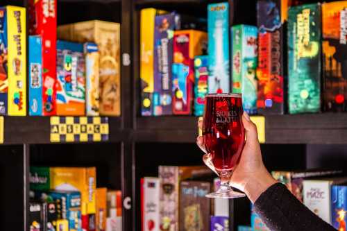 A hand holds a glass of red drink in front of a colorful shelf filled with board games.