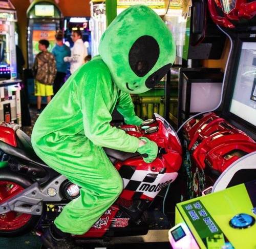 A person in a green alien costume rides a motorcycle arcade game in an amusement arcade.