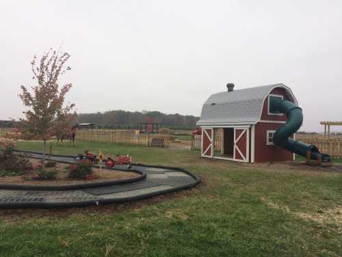 A playground featuring a red barn with a slide, a winding path, and a grassy area with trees in the background.