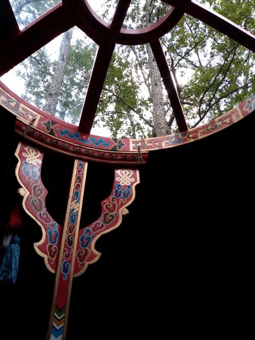 View from inside a yurt, showcasing colorful wooden beams and a circular opening revealing trees above.