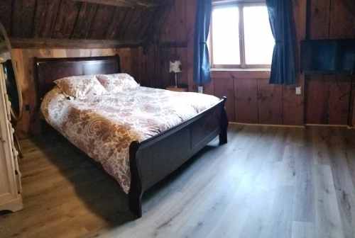 Cozy bedroom with a wooden bed, patterned bedding, and a window, featuring rustic walls and a TV on the side.