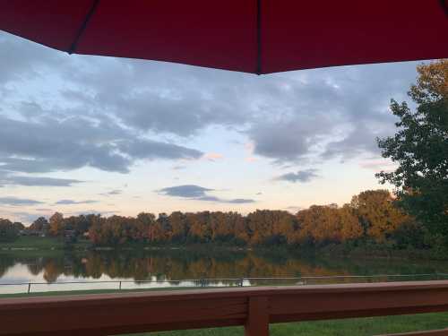 A serene lake view under a red umbrella, with trees and clouds reflecting on the water at sunset.