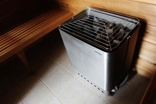 A sauna heater with a metal casing and rocks, positioned next to a wooden bench in a dimly lit sauna.