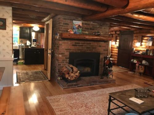 Cozy cabin interior with wooden beams, a brick fireplace, and a view of the kitchen and dining area.