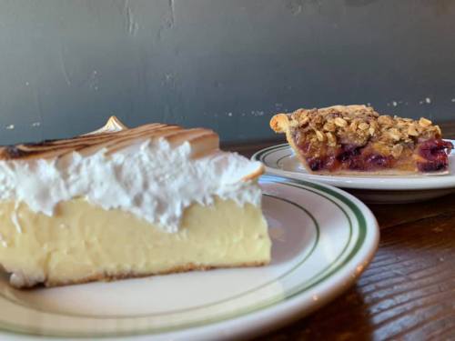 A slice of creamy dessert topped with whipped cream next to a slice of fruit pie with a crumbly topping on a plate.