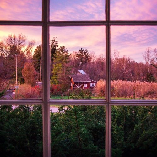 View through a window showcasing a rustic red barn surrounded by trees and colorful autumn foliage at sunset.