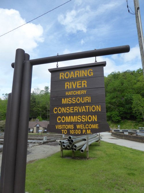 Sign for Roaring River Hatchery, Missouri Conservation Commission, welcoming visitors until 10:00 PM. Green hills in background.