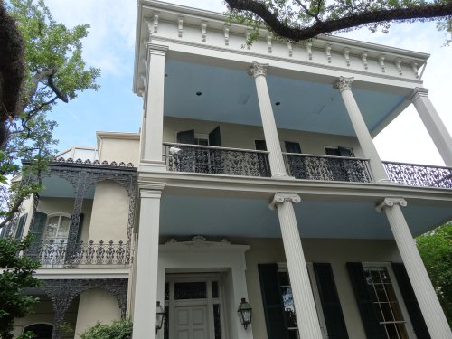 A historic two-story house featuring columns, balconies, and intricate ironwork, surrounded by greenery.