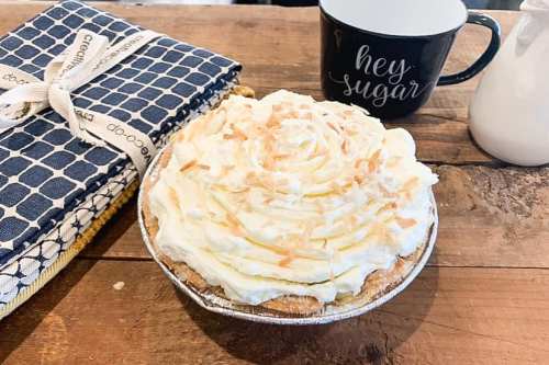 A coconut cream pie with whipped topping on a wooden table, alongside a black mug and a patterned kitchen towel.