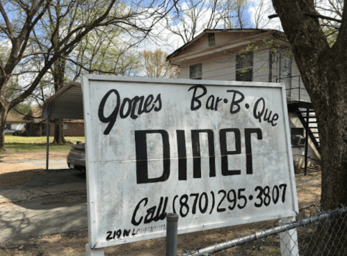 Sign for Jones Bar-B-Que Diner with contact number, set in a rural area with trees and a building in the background.