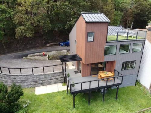 A modern two-story house with a wooden exterior, large windows, and a deck, surrounded by greenery and a winding road.
