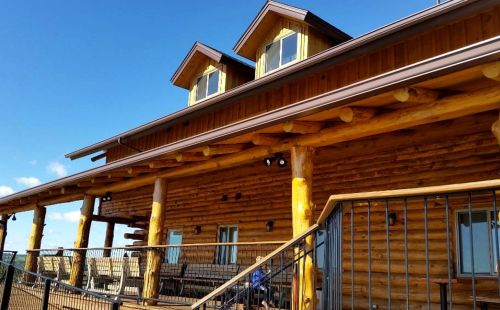 A rustic log cabin with a large porch, featuring wooden beams and a clear blue sky in the background.