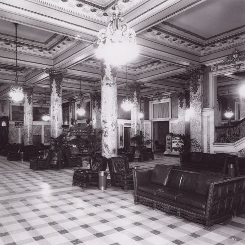 A spacious, elegant lobby with marble columns, vintage furniture, and decorative lighting, featuring a checkered floor.