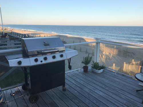 A grill on a wooden deck overlooking a sandy beach and ocean under a clear sky.