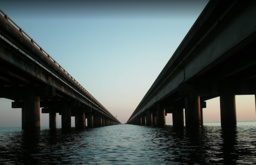A view of two parallel bridges extending over calm water, with a soft sunset glow in the background.